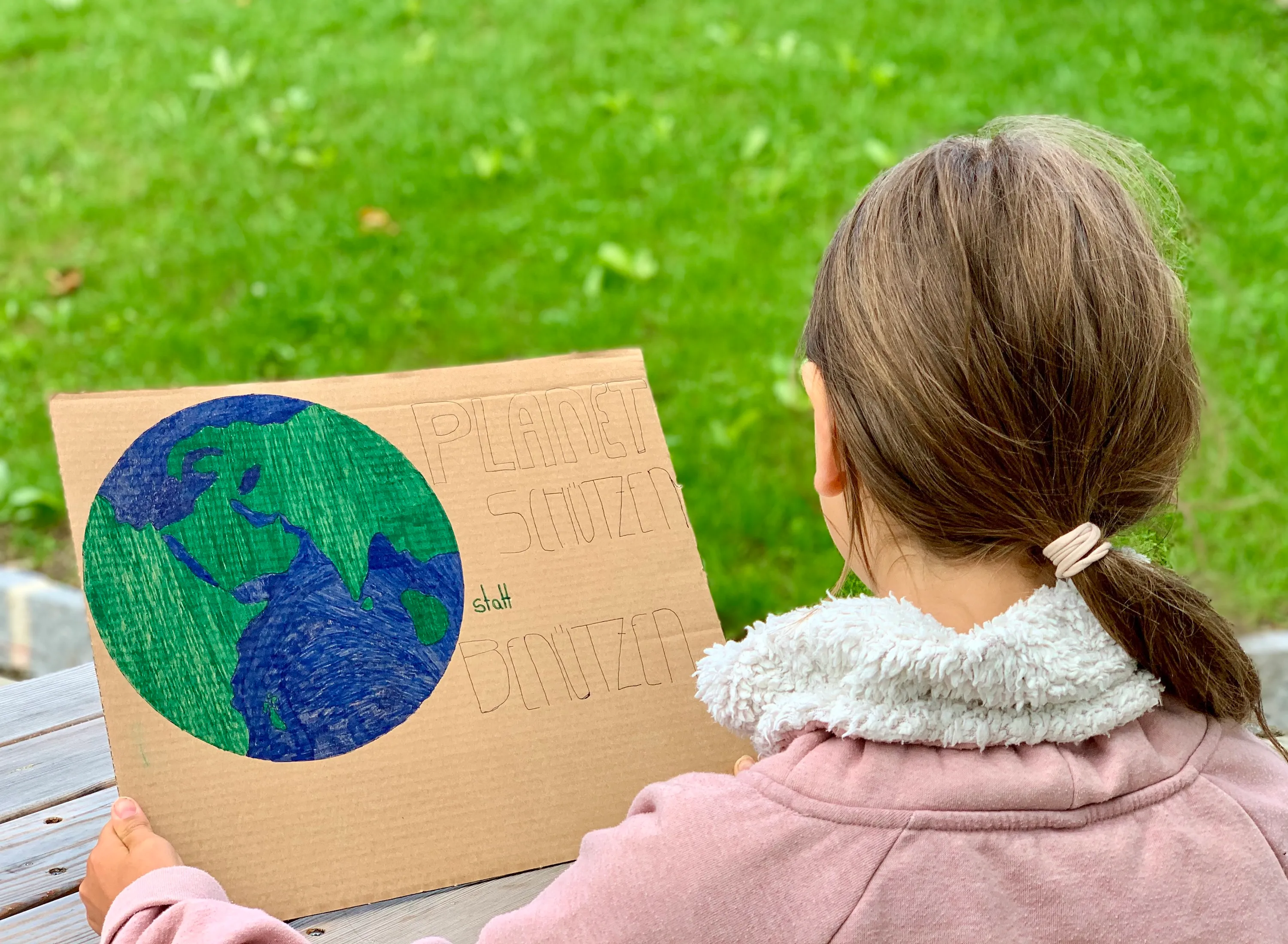 Ein Kind mit langen Haaren ist von hinten zu sehen und betrachtet ein Demo-Plakat mit einer Zeichnung der Erde und der Aufschrift: "Planeten schützen statt benützen!"